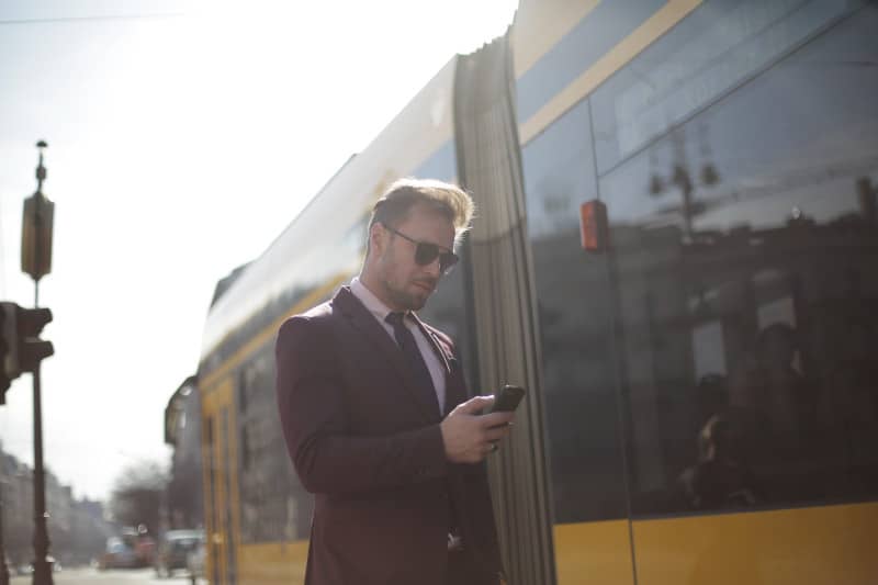 Ein Geschäftsmann benutzt ein mobiles Gerät unterwegs, um Mobilität zu verdeutlichen, während im Hintergrund eine Straßenbahn vorbeifährt.