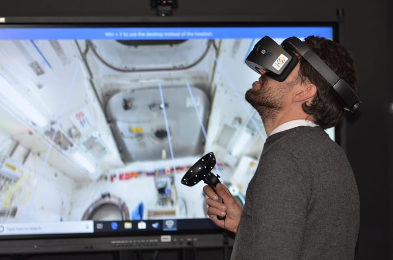 Ein Mann mit einer aufgesetzten VR-Brille erkundet die Weltraumstadion ISS. Seine Sicht ist auf dem Bildschirm hinter ihm sichtbar.